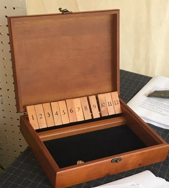 471468079_10160616376157256_5999015292391894835_n - Luzerne County Historical Society Open wooden box with a row of numbered wooden tiles 1–12 inside, black felt-lined bottom, and a small die visible in the tray.