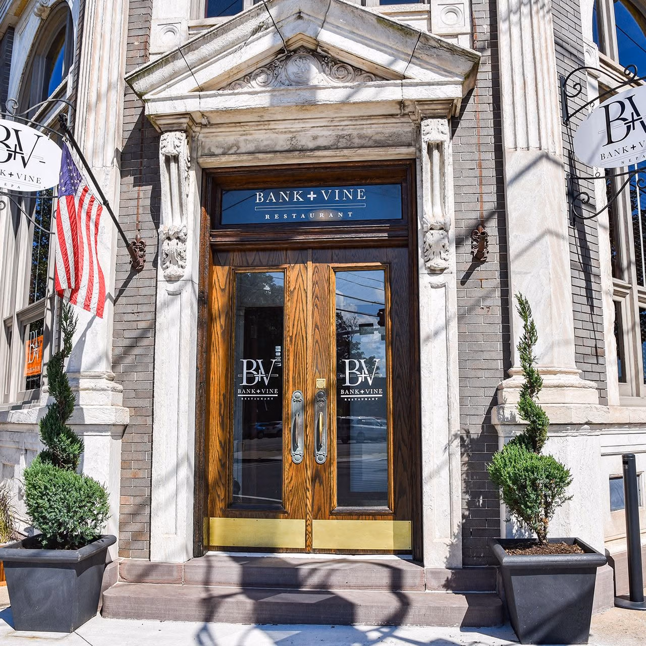 Front entrance of Bank+Vine Restaurant with wooden double doors and blue sign above reading 'BANK+VINE RESTAURANT' on a brick façade, with potted evergreens and an American flag nearby.