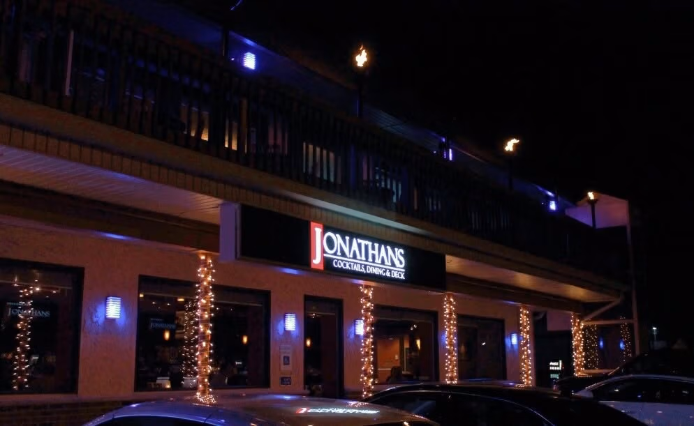 Night exterior of Jonathan's Cocktail, Dining & Deck illuminated with string lights around the columns and patio area.